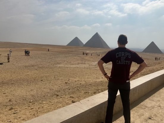 Person standing in front of the Great Pyramids of Giza with hands on their hips, wearing a shirt labeled 'Corps of Cadets'.