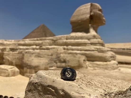A ring rests on a stone in the foreground with the Great Sphinx of Giza and a pyramid in the background under a clear sky.