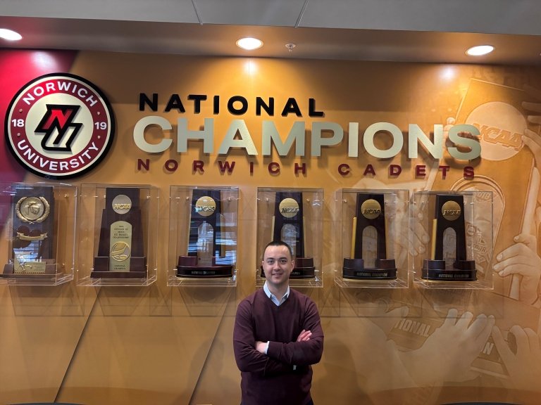 An individual standing in front of a trophy display titled "National Champions Norwich Cadets" with various awards encased in glass shelves.