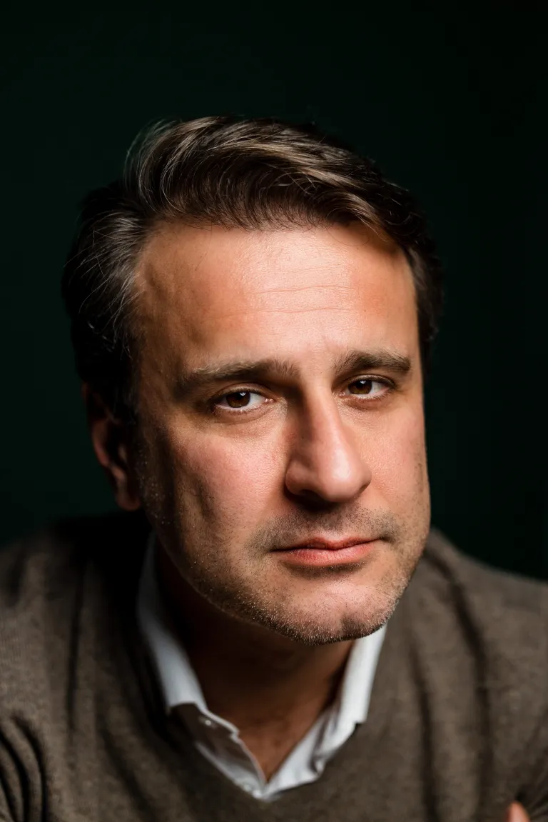 Person in a collared shirt and sweater looks toward the camera against a dark background.