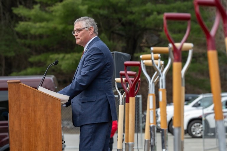 Phil Sussman stands behind the podium during the groundbreaking with a line of shovels in the dirt behind him.