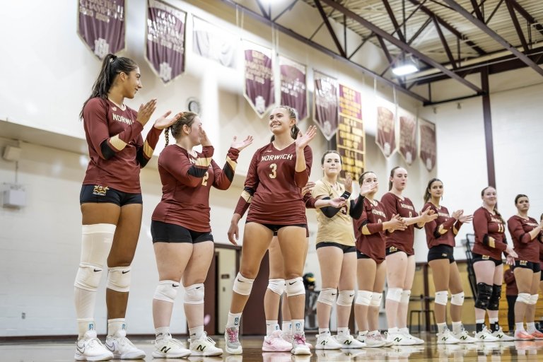 Rae Lawing in her volleyball uniform walks in front of a line of teammates in the gym.