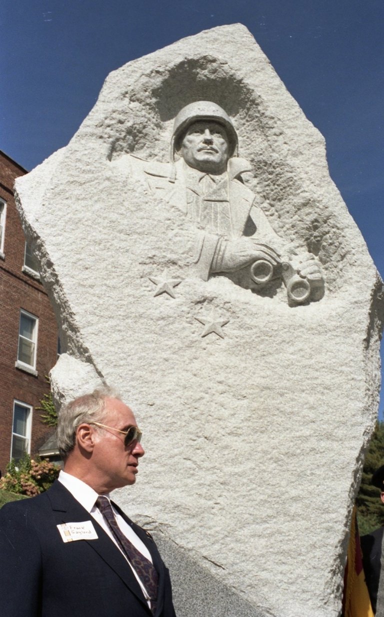 A close-up shot in portrait. Frank Gaylord stands in sunglasses and black suit and tie, with nametag sticker on chest. He is just next to the statue of Harmon.