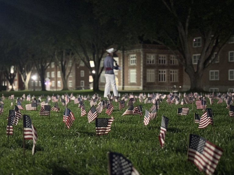 A person in a military uniform at a memorial with numerous American flags planted in a grassy field, with a classic brick building in the background illuminated by warm lights at dusk.