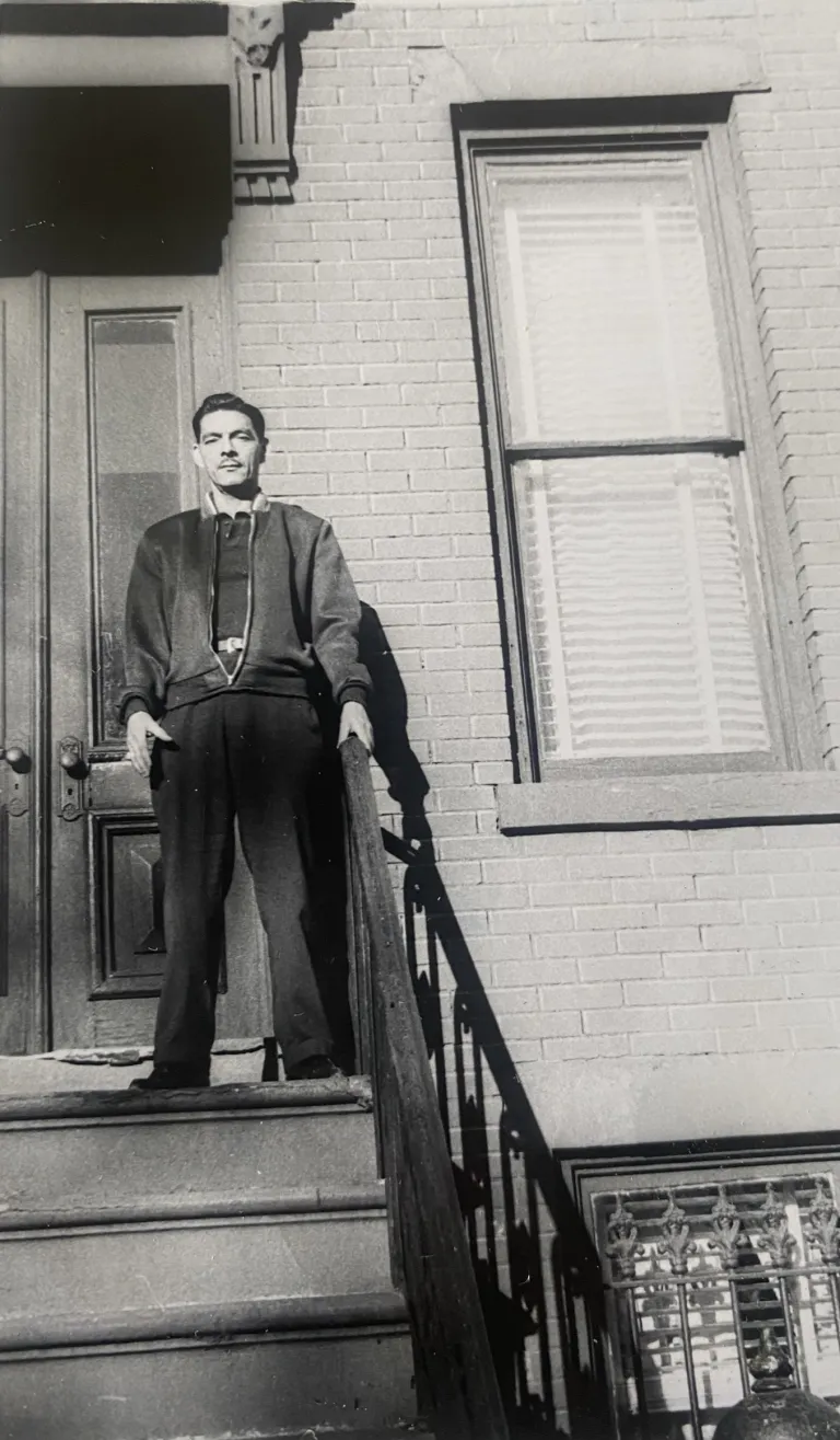 Jose Barreno in a black and white photo, standing in slacks and jacket holding the rail on the stairs of a brick New York building.