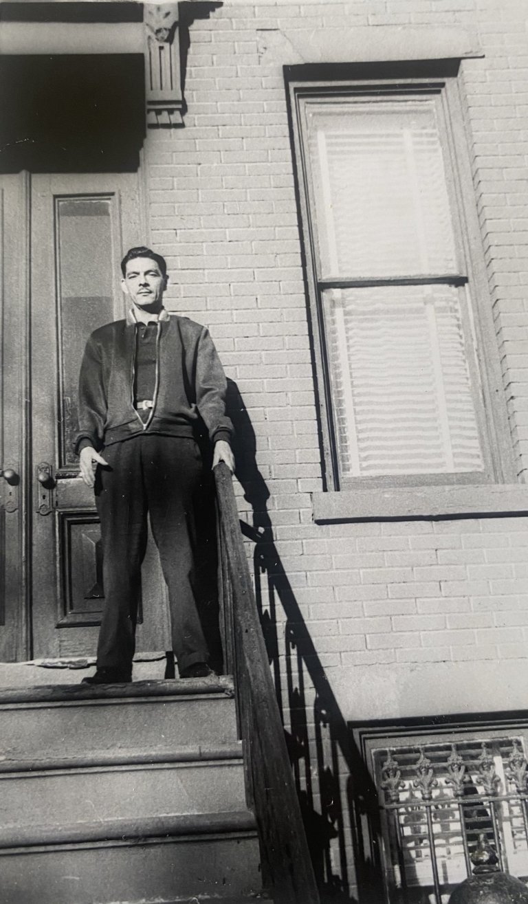 Jose Barreno in a black and white photo, standing in slacks and jacket holding the rail on the stairs of a brick New York building.