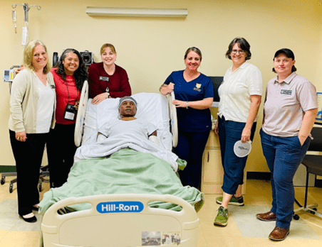 (Left to Right) Jennifer Coutu, Rea Colby, Janie Szalajeski-Hull, Amy Martone, Olga Ryan, and Cindy Freudenthal pose with a high-fidelity 3G manikin, Thomas, in one of the acute care hospital rooms located in the School of Nursing Clinical Simulation Center.