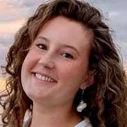 Woman with curly brown hair, white earrings, and a white top standing outdoors at sunset.