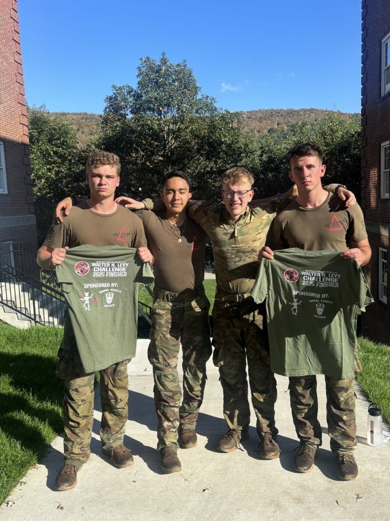 Four individuals in military-style uniforms standing outdoors between two brick buildings. They are posing together with arms around each other. Two of them are holding green shirts that read “Walter N. Levy Challenge 2025 Finisher." The background features trees and hills under a clear blue sky.