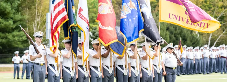 A group of cadets march while holding an American flag and one for each branch, and the University's flag.