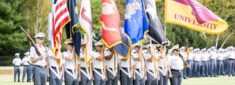 A group of cadets march while holding an American flag and one for each branch, and the University's flag.