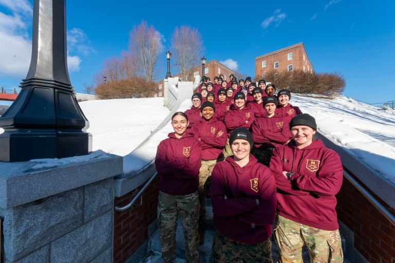 Group in maroon hoodies and camouflage pants stands on snowy outdoor steps posing for a photo.