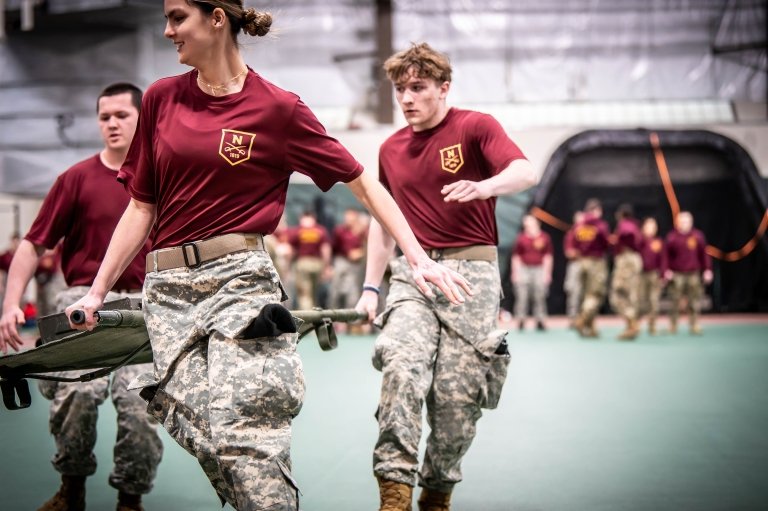 People in maroon shirts and camouflage pants run indoors carrying a stretcher together.
