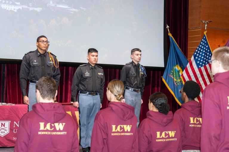 Three people in dark uniforms stand on stage before others in maroon LCW Norwich University hoodies.