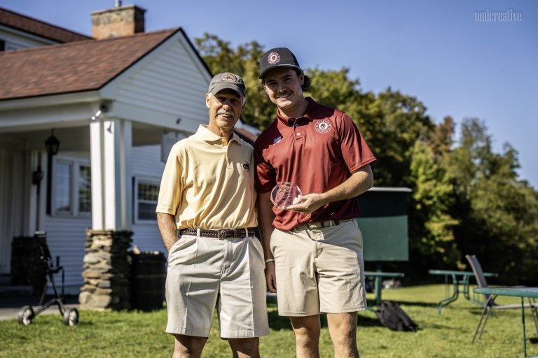 Two people in golf attire stand outside near a white building, one holding a clear award.