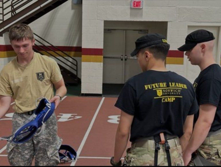 Three cadets stand on an indoor track, two wearing black shirts that read “Future Leader Camp, Norwich University,” and one holding climbing gear in a tan shirt with camouflage pants.