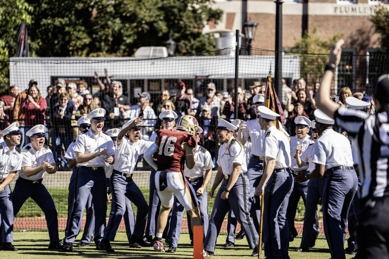 Football player in a maroon uniform celebrates a touchdown with cheering cadets in white uniforms and hats near the end zone.