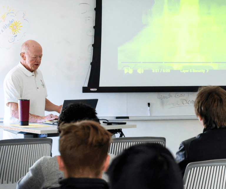 A professor lectures to a class in front of projection screen.
