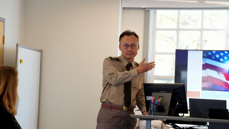 A man in a khaki military-style uniform is standing at the front of a classroom, gesturing with one hand while speaking. Behind him, a presentation slide is visible on a screen, featuring part of the American flag. A woman seated in the foreground is listening attentively. The setting appears to be an academic or professional presentation, possibly related to military or international affairs.