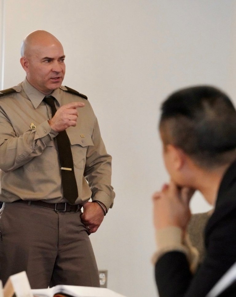 A man in a khaki military-style uniform is standing and gesturing while speaking to someone seated in the foreground. His posture and expression suggest he’s making a point or giving instructions. The person listening is blurred and appears attentive, with hands resting under their chin. The setting looks like a classroom or seminar space, with papers and notebooks visible on a table.