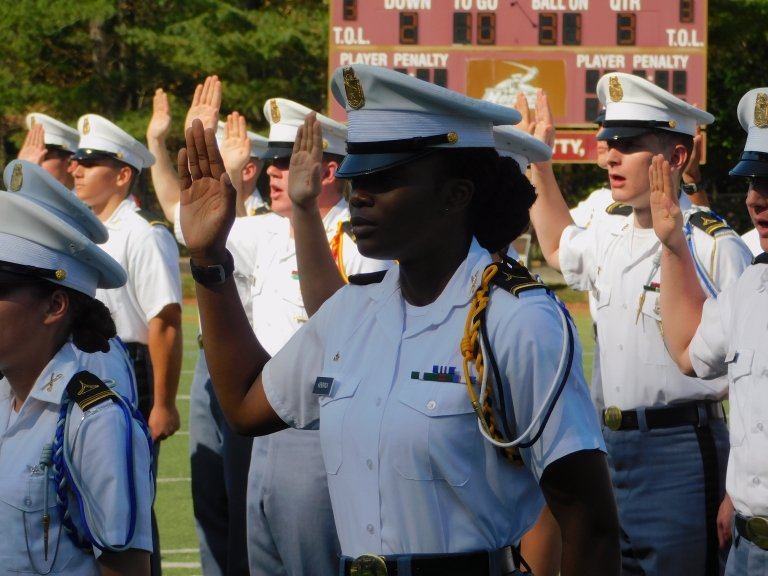 Cadets in uniform stand on a field with raised hands during a formal ceremony.