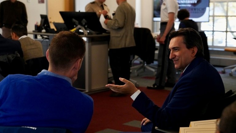 Two men are engaged in conversation in a professional setting, with one gesturing while speaking and the other listening attentively. Both are seated among others in a room that appears to host a meeting or seminar. In the background, several people stand near a desk and computer station, talking in small groups. The atmosphere feels collegial and focused, suggesting a formal or academic gathering.