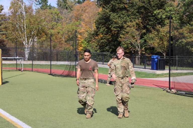 Two individuals in military fatigues walking on a turf athletic field. One is wearing a brown T-shirt, while the other is in full camouflage uniform. The setting appears to be an outdoor training or drill exercise, with trees showing fall foliage in the background and a running track visible along the field.