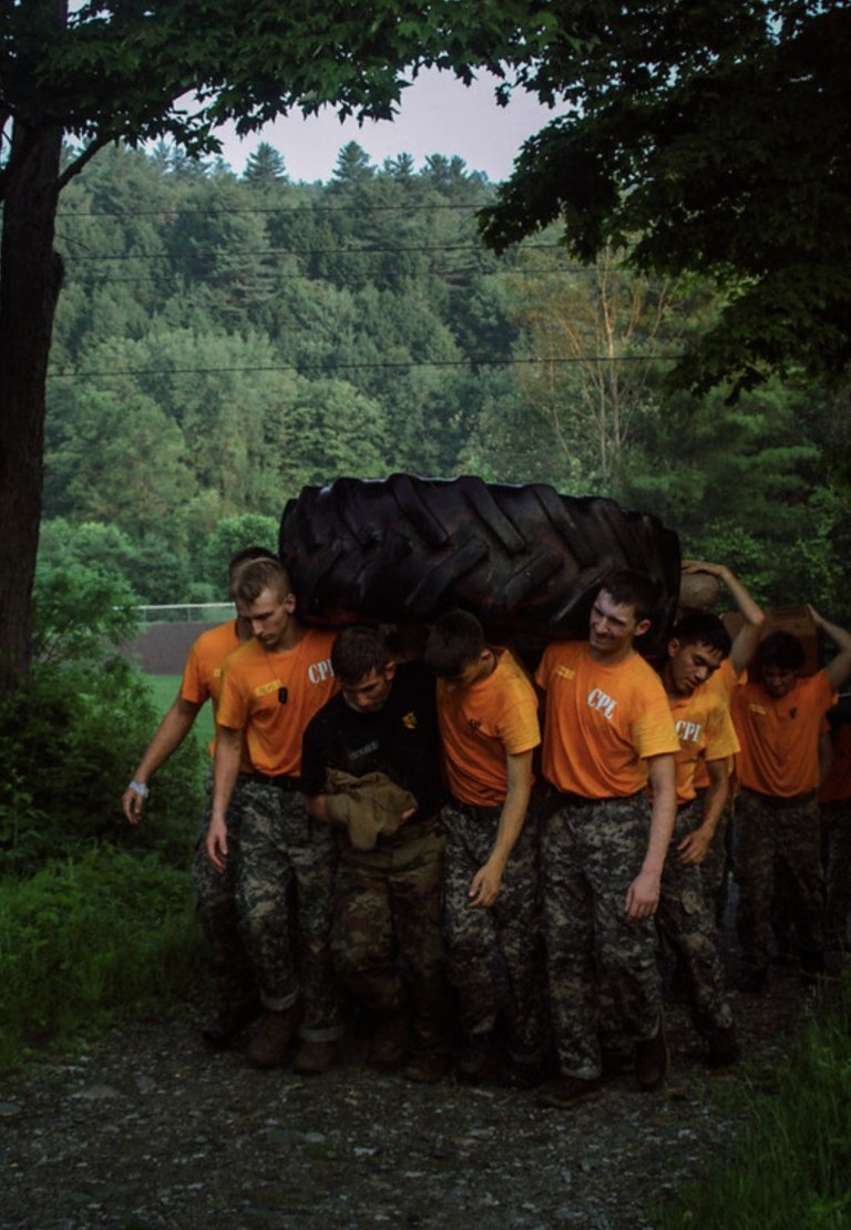 A group of cadets in orange shirts and camouflage pants work together to carry a large tractor tire along a wooded path.