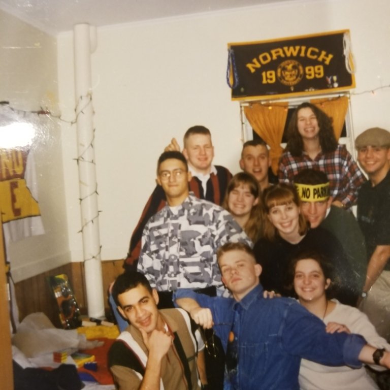 A group of young adults is gathered closely together, smiling and posing for a fun, casual photo inside a dorm-style room. A Norwich University banner marked “1999” hangs above a small window decorated with string lights. The individuals are dressed in a variety of 1990s-style clothing — including flannel shirts, denim, camouflage patterns, and a “No Parking” headband — giving the scene a lively and nostalgic atmosphere typical of a college social gathering from that era.