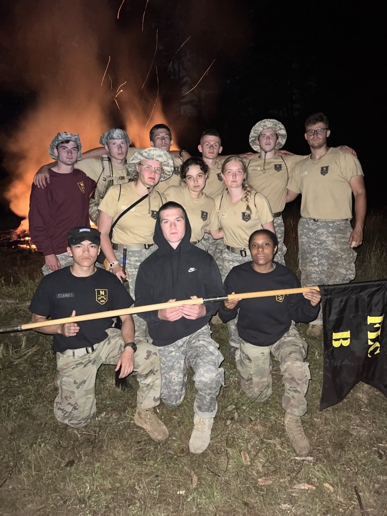 A group of cadets take a group photo with a bonfire in the background.