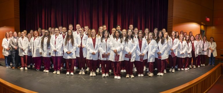 A large group of nursing students stand on a stage wearing white coats and maroon scrubs, posing for a formal group photo.