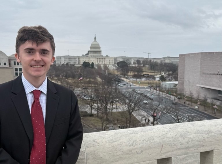 A student in a black suit stands on the Speaker's Balcony with D.C. behind him.