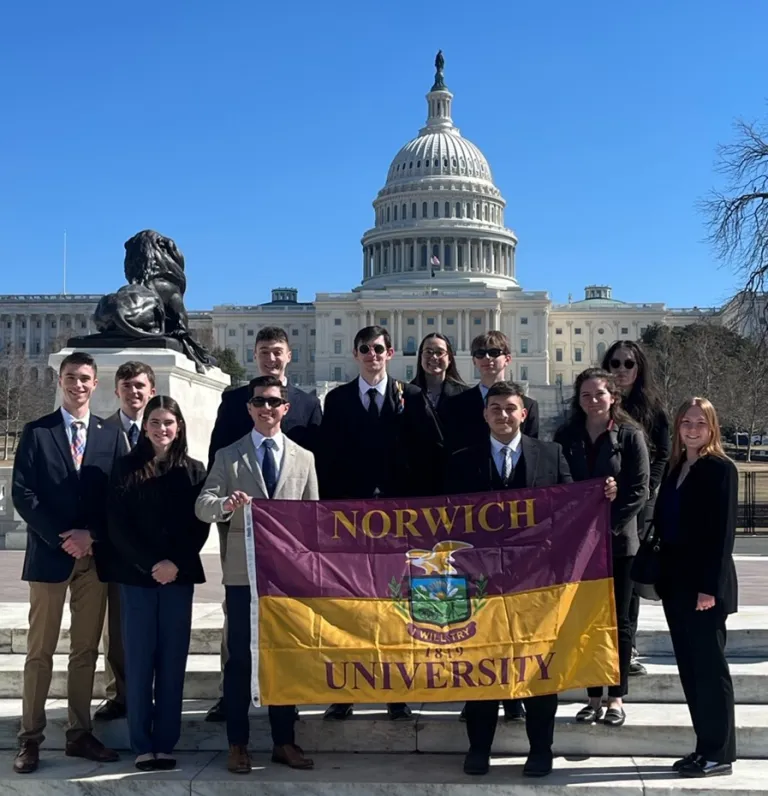 A group of students stand on the stairs of the Capitol holding a Norwich University flag.