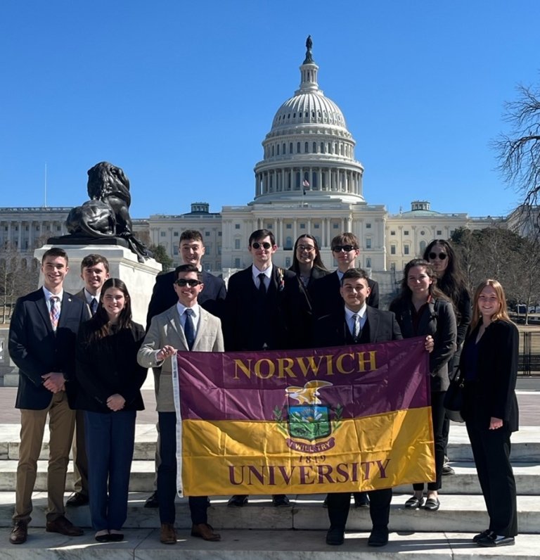 A group of students stand on the stairs of the Capitol holding a Norwich University flag.