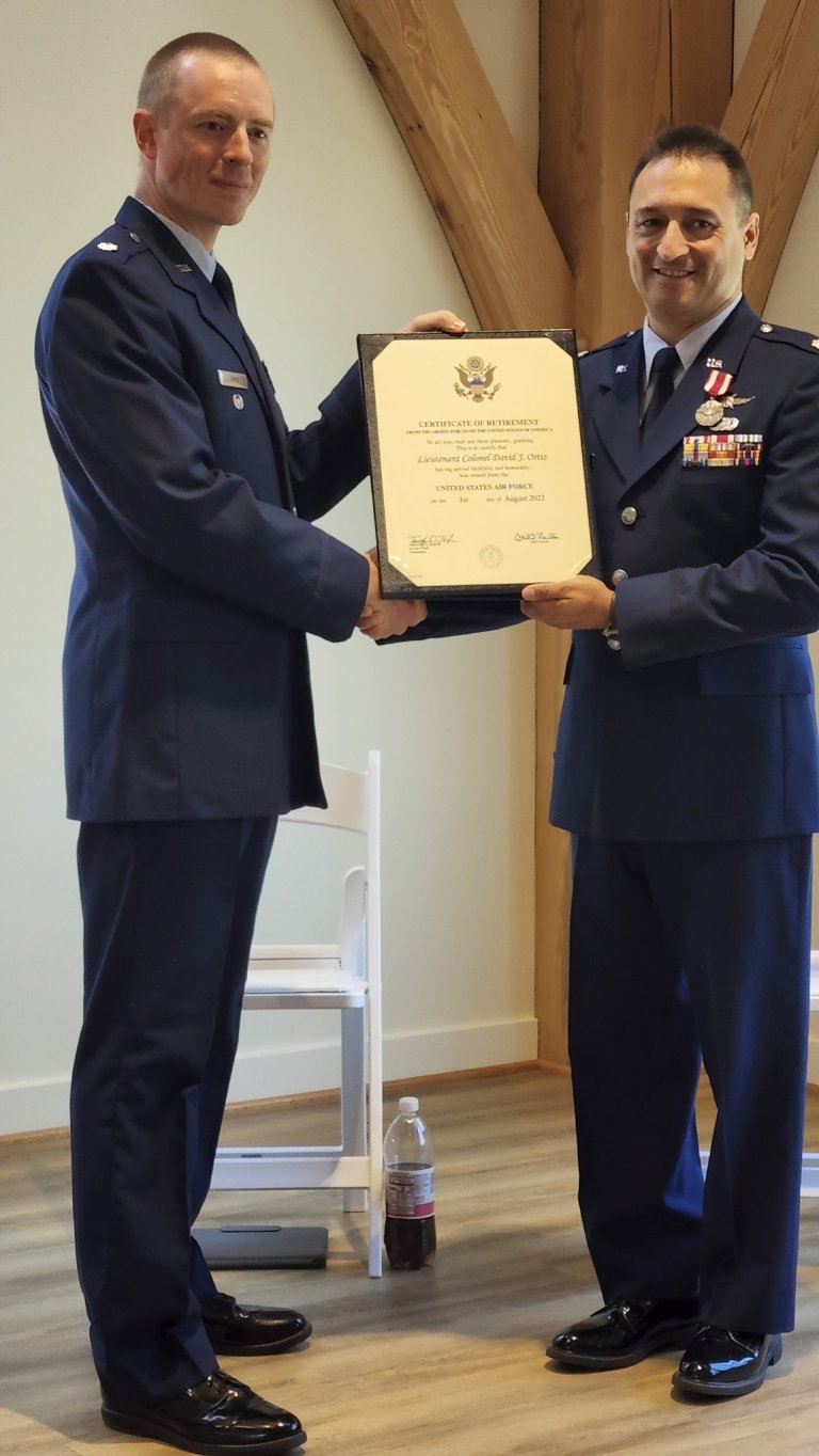 Two U.S. Air Force officers stand side by side indoors, each dressed in formal blue service uniforms. They’re smiling as they hold a framed Certificate of Retirement together, marking a ceremonial moment for one of them. The setting has a simple wooden-beam backdrop and light-colored walls, adding a modest, professional tone to the occasion.