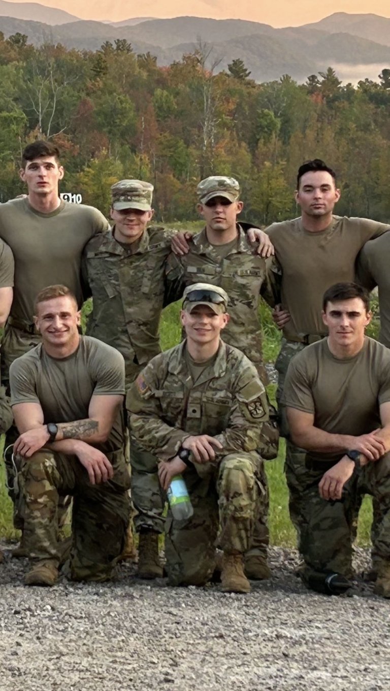 A group of soldiers in camouflage uniforms and tan shirts pose outdoors with a scenic background of trees and mountains at sunset.