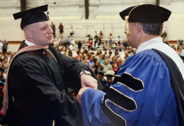 A man smiles as he shakes a hand a receives his diploma on stage.
