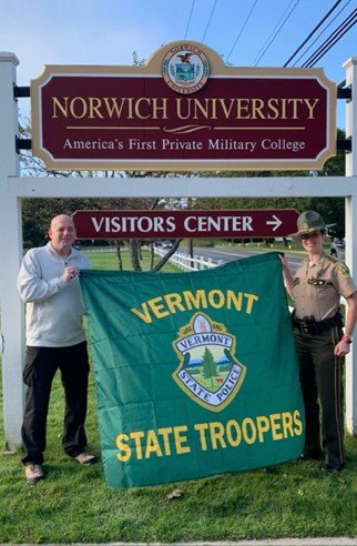 A man and Vermont state trooper hold a Vermont State Troopers flag under the Norwich University campus sign.