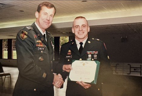 Two men in military uniforms smile and shake hands, as the man on the left hands a diploma to the one on the right.