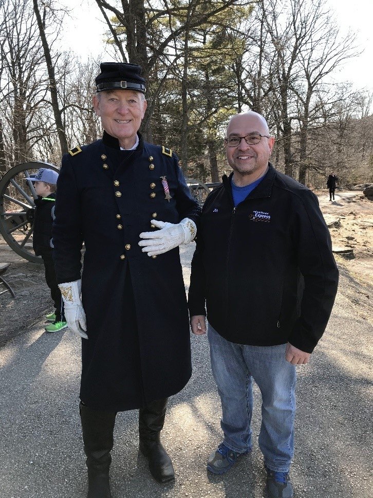 A smiling man stands next to another who is dressed in historic American military attire.