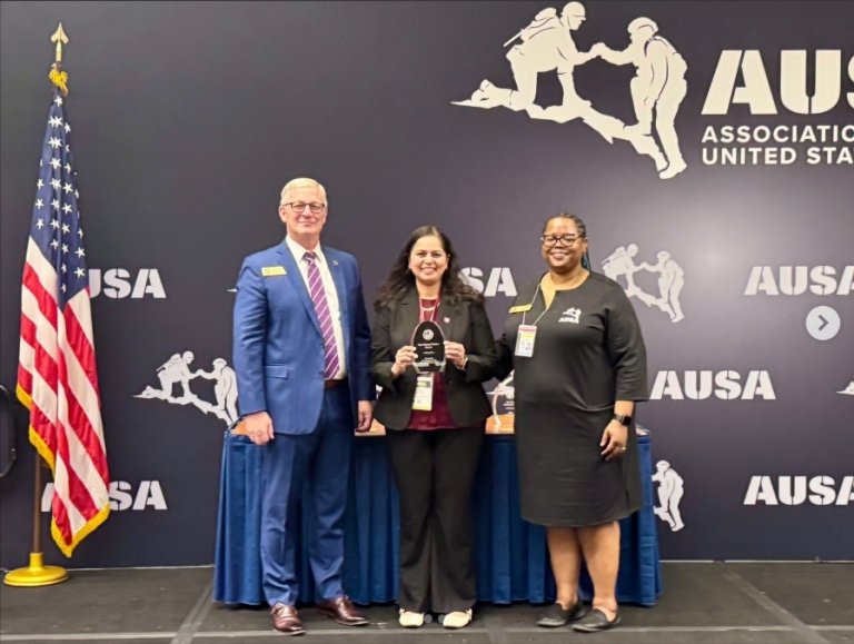 A group of three pose for a photo, with the woman in the center holding a plaque.