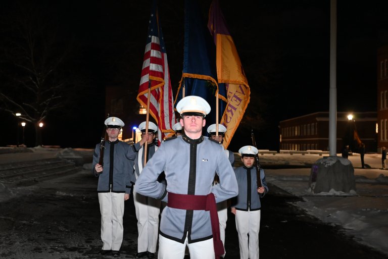 A group of cadets stand outside at night with the colors.