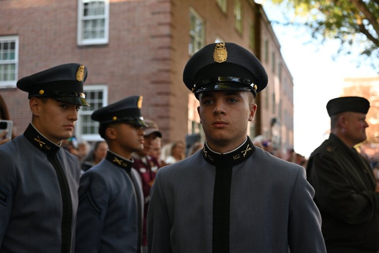 Cadets in gray dress uniforms and black caps stand in formation on a street lined with brick buildings and onlookers.