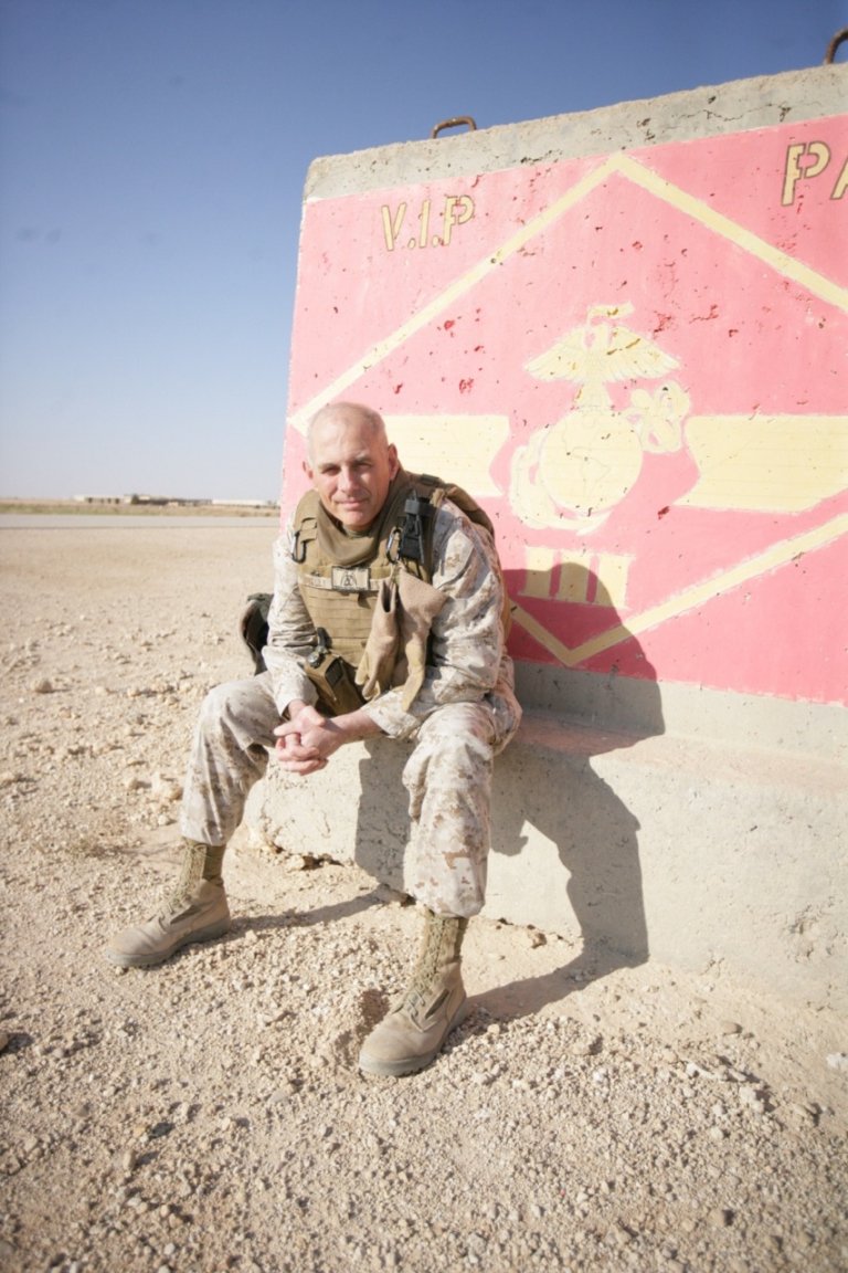 Person wearing desert camouflage and body armor while sitting against a concrete barrier painted with a Marine Corps insignia.