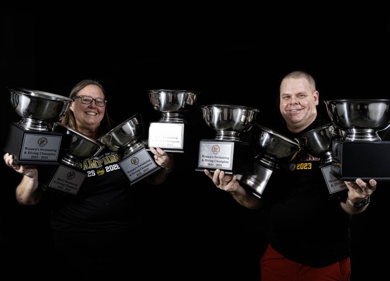 Two people in dark shirts hold multiple large silver trophies against a black background.