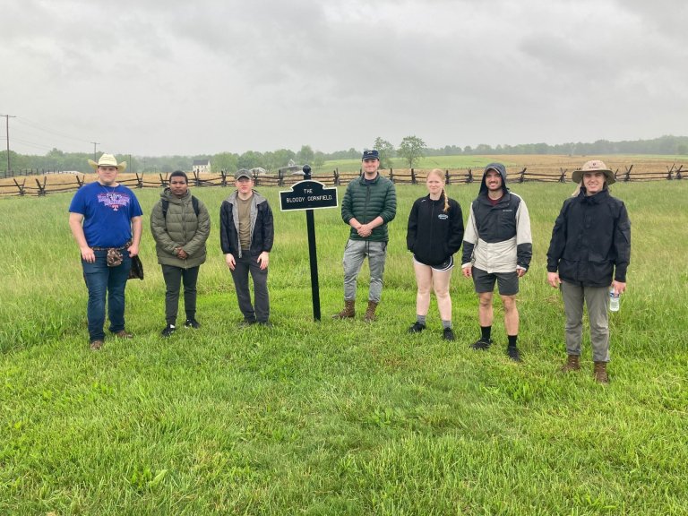 Seven people stand in a grassy field near a sign that reads “The Bloody Cornfield” with a split‑rail fence and cloudy sky behind them.