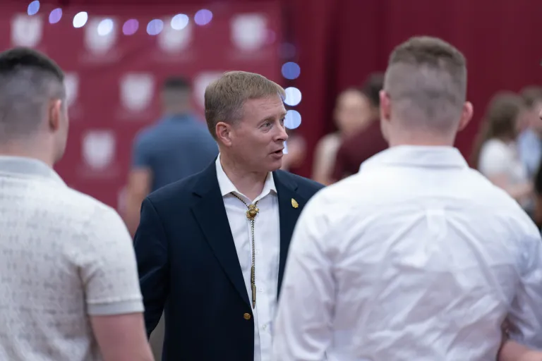 A person in a dark blazer with a bolo tie speaks with two people in white shirts at an indoor event with red banners in the background.