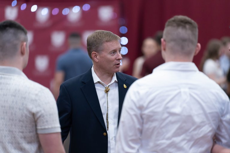 A person in a dark blazer with a bolo tie speaks with two people in white shirts at an indoor event with red banners in the background.
