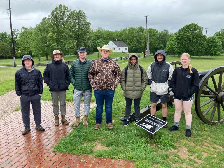 Seven people stand on a wet brick path beside a cannon in a grassy field with trees and a small white house in the background.