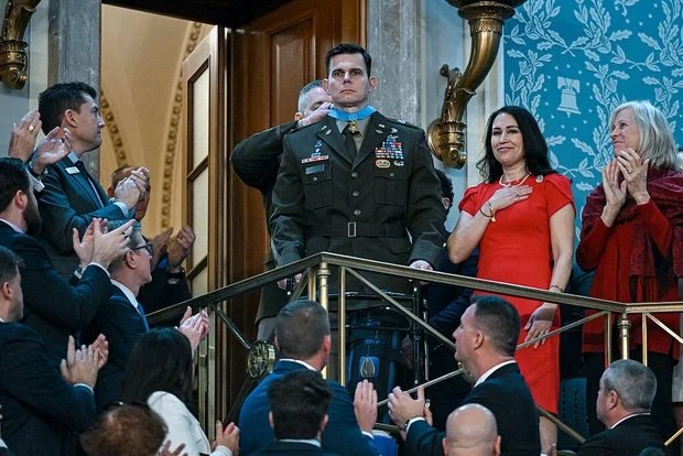  A person in a decorated military uniform stands on a balcony as people around them applaud in a large hall.
